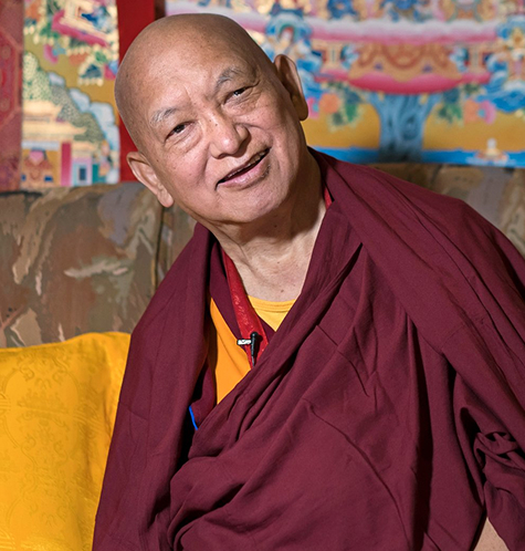 A smiling monk wearing traditional robes sits on a couch, surrounded by colorful Tibetan artwork.
