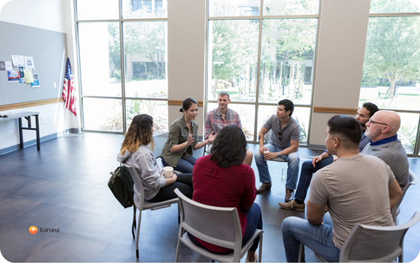 A group of eight people sits in a circle, engaged in discussion, with large windows and greenery in the background.