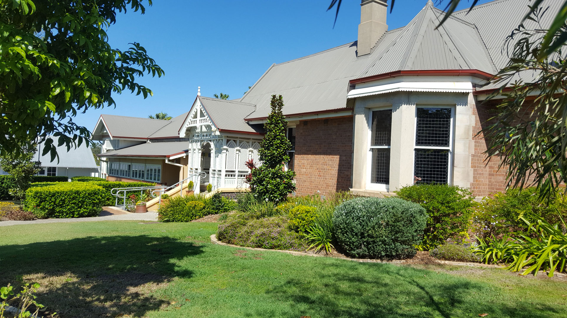 A well-maintained garden surrounds a brick house with a decorative porch and multiple green shrubs under a clear blue sky.