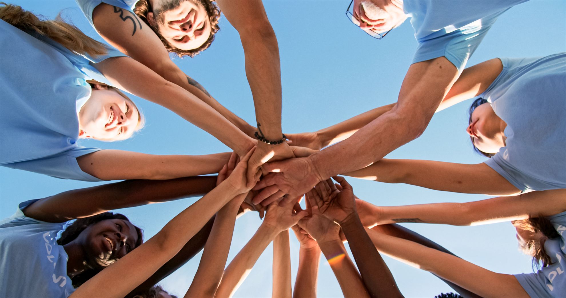 A group of diverse individuals, wearing blue shirts, join hands together in a circle against a clear blue sky.