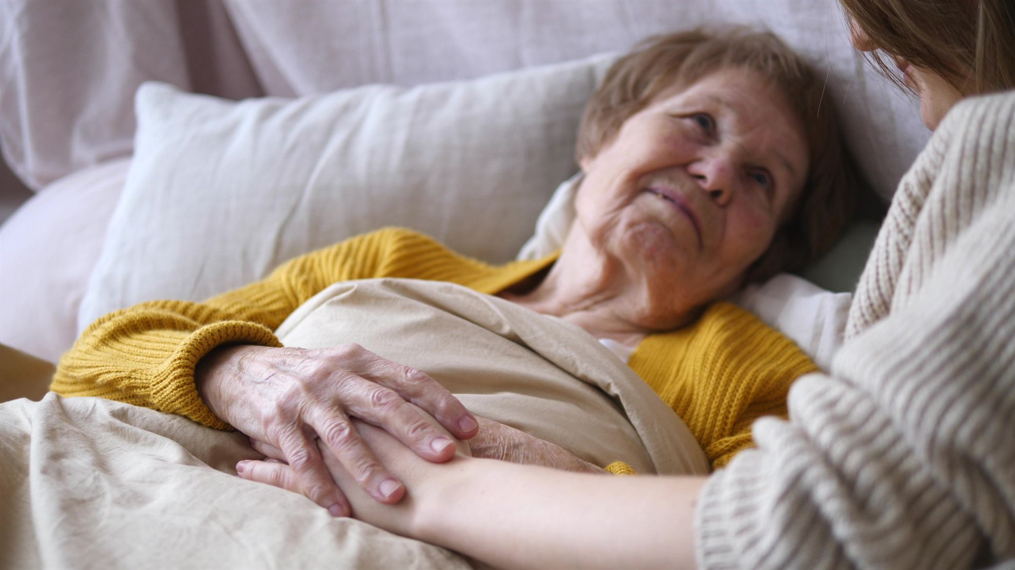 An elderly woman in bed smiles at a younger person holding her hand, both are wrapped in blankets.