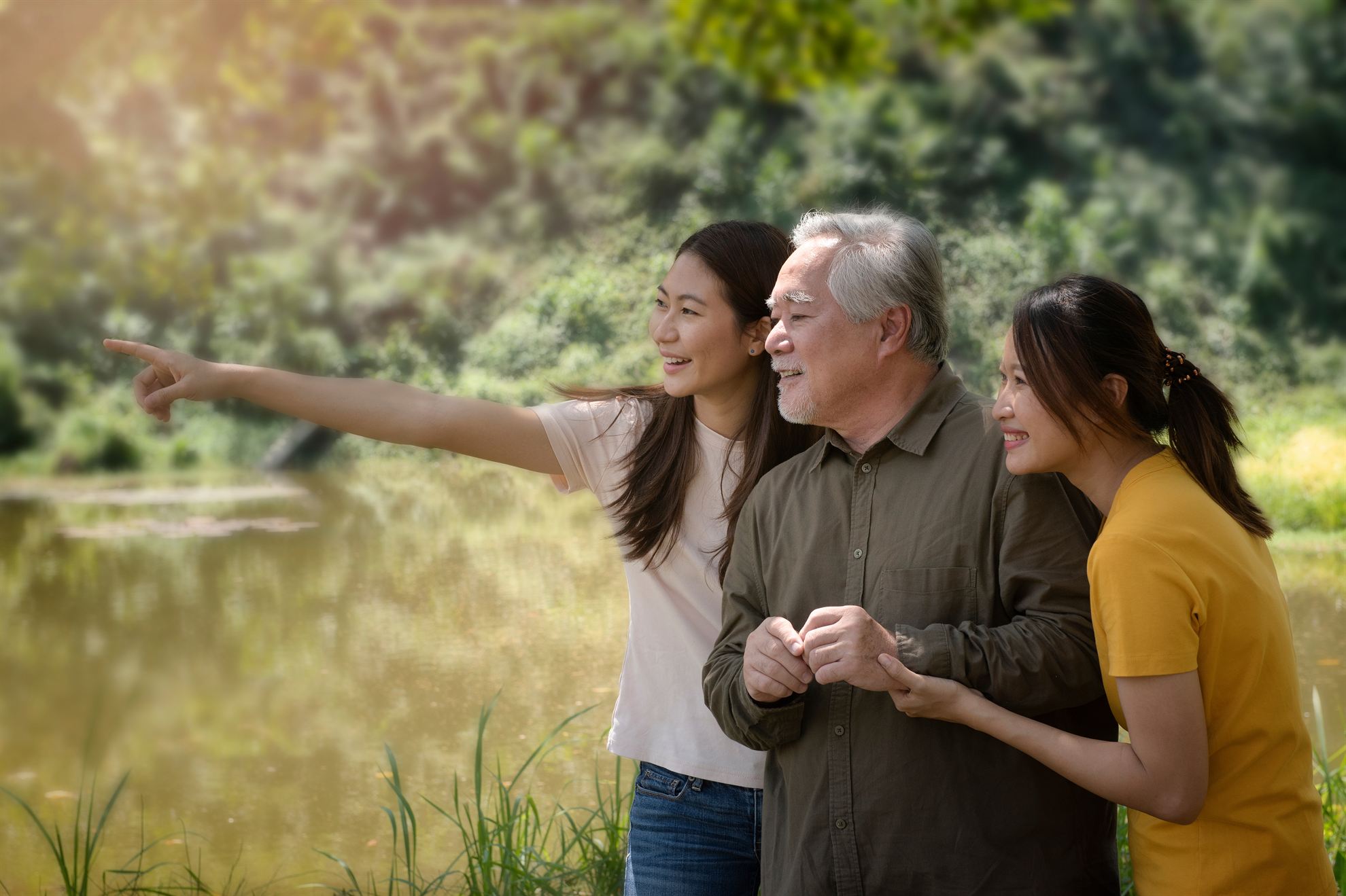 Two women and a man stand by a pond, smiling and looking towards the distance, surrounded by greenery.