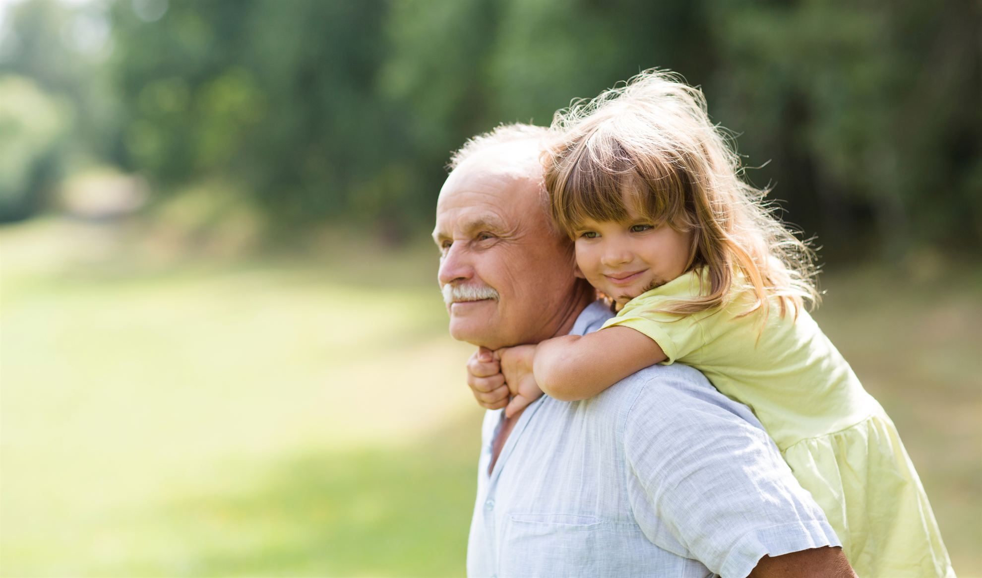 An elderly man carries a young girl on his back, both smiling, in a green outdoor setting.