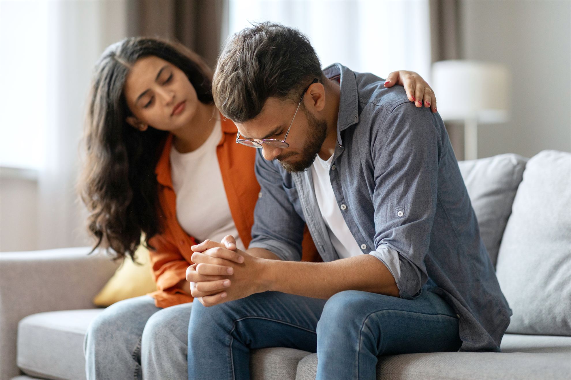 A woman gently comforts a man sitting on a couch; he looks down, appearing distressed. Natural light fills the room.