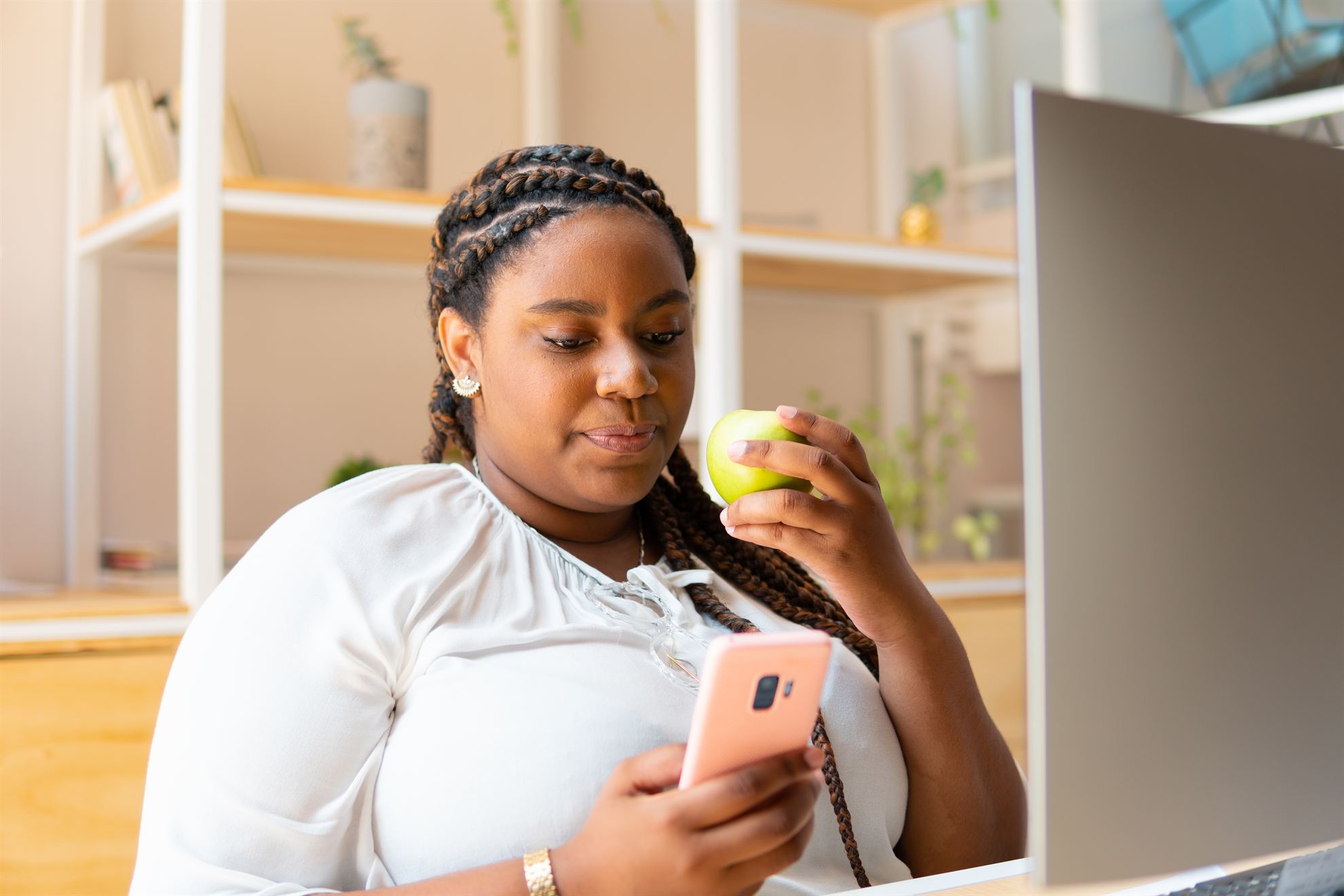 Woman with braided hair enjoying a green apple while using a smartphone in a bright office setting.