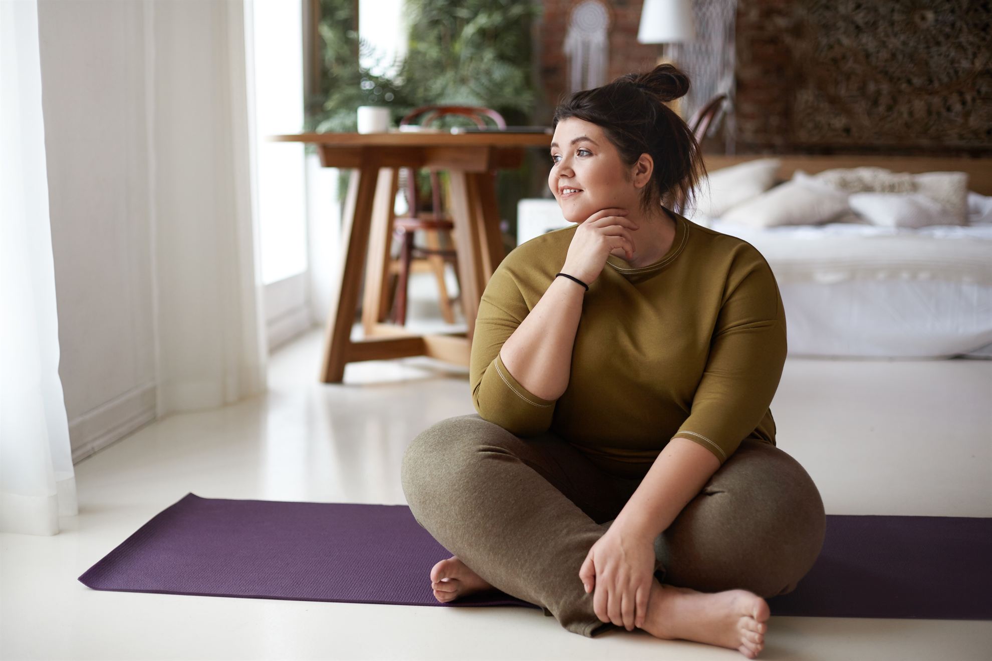 A woman sits cross-legged on a yoga mat, smiling, in a bright, cozy room with plants and a wooden table.
