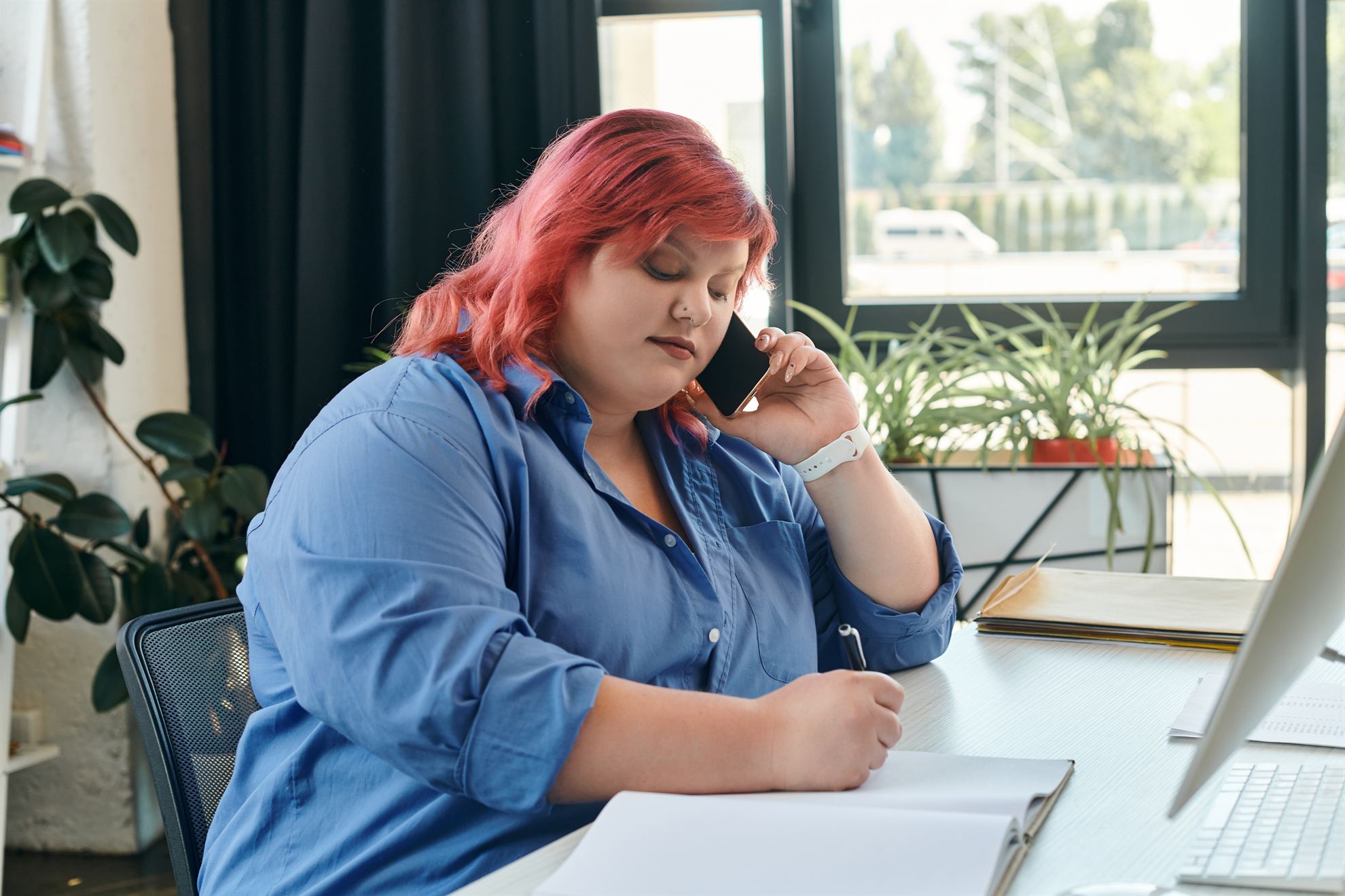A woman with pink hair sits at a desk, holding a phone to her ear and writing in a notebook.
