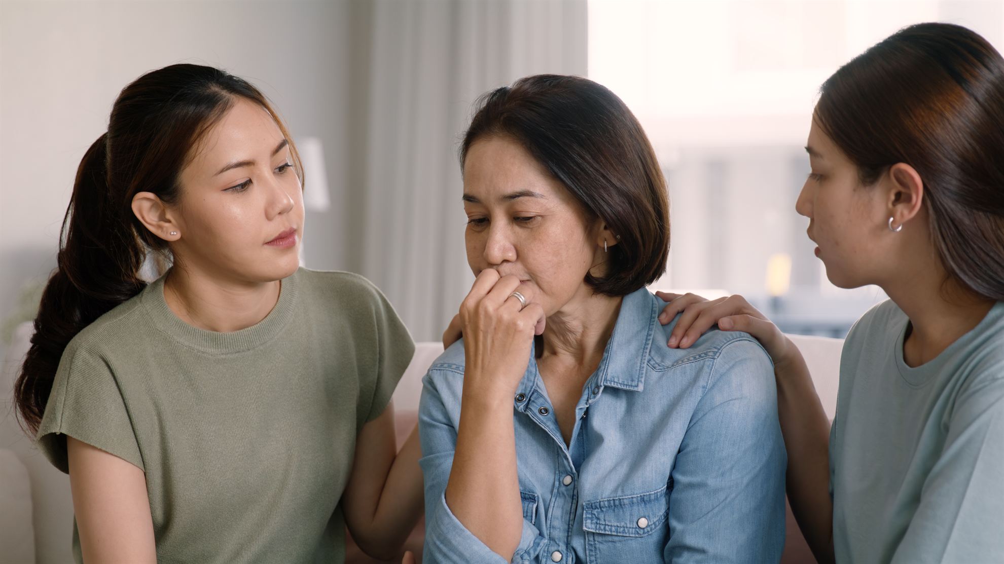 Two women comfort an older woman who appears distressed, sitting together in a softly lit indoor setting.