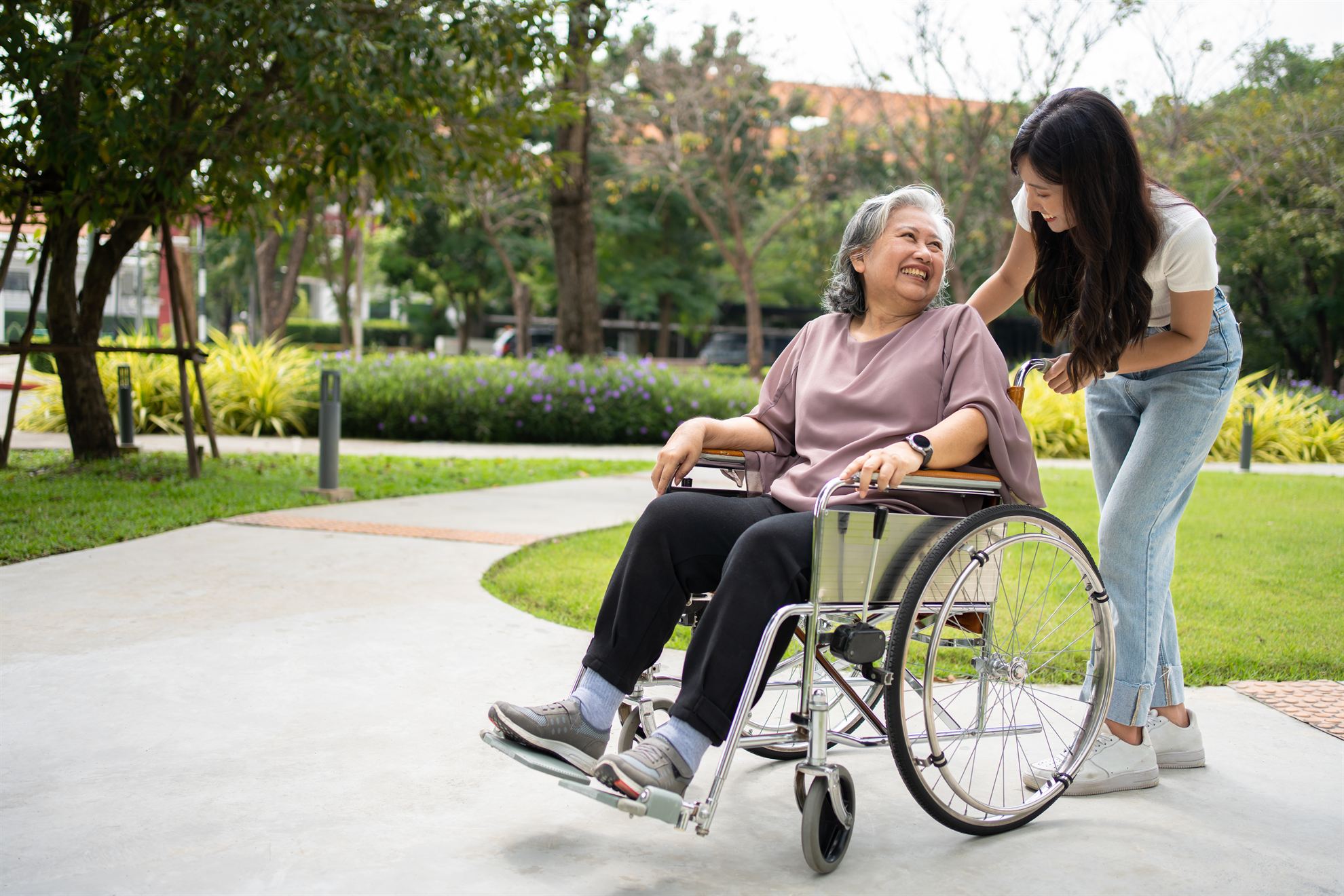 An older woman in a wheelchair smiles at a young woman standing beside her in a sunny park.