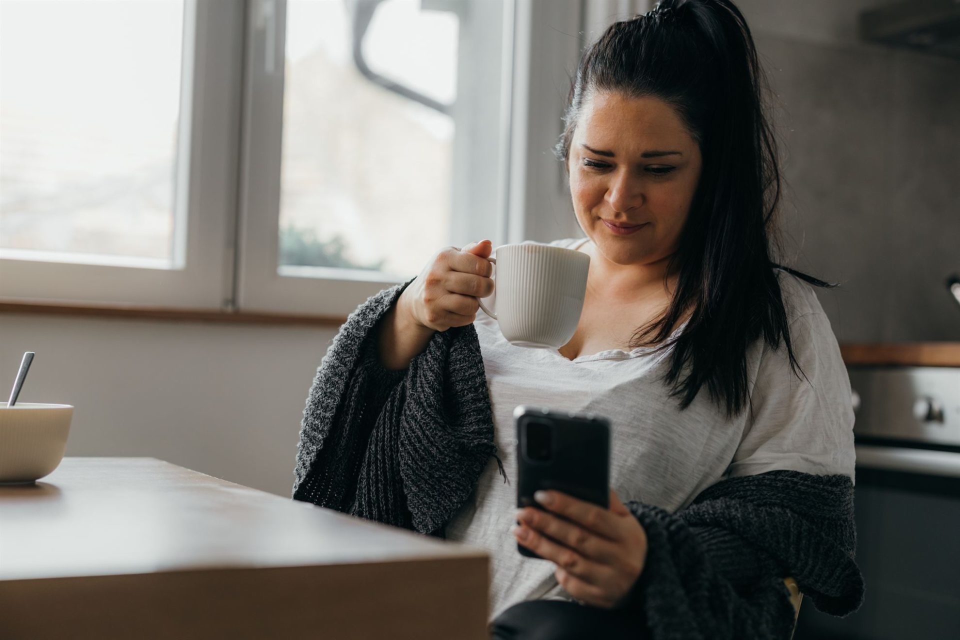 A woman in a cozy sweater sits at a table, holding a cup and looking at her phone. Natural light enters through a window.
