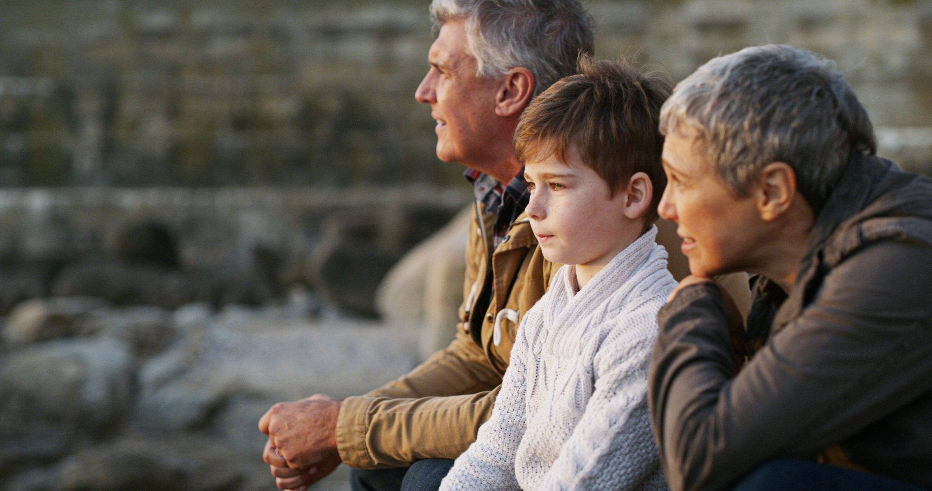 A man, a woman, and a boy sit together on rocks, gazing into the distance, with a blurred background.