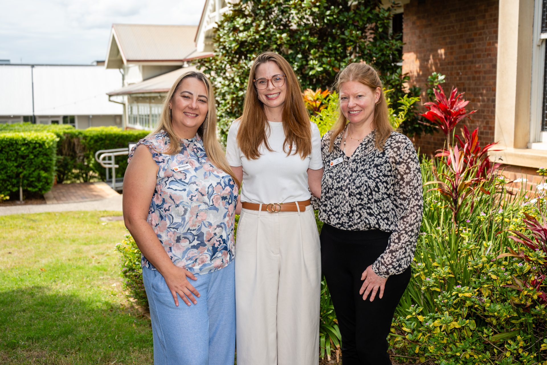Three women stand together outdoors, smiling, with greenery and flowers in the background.