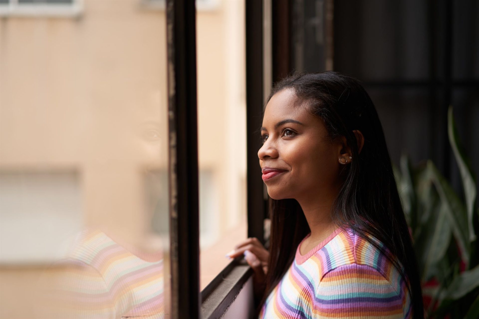 A woman with long dark hair gazes thoughtfully out of a window, wearing a colorful striped sweater.