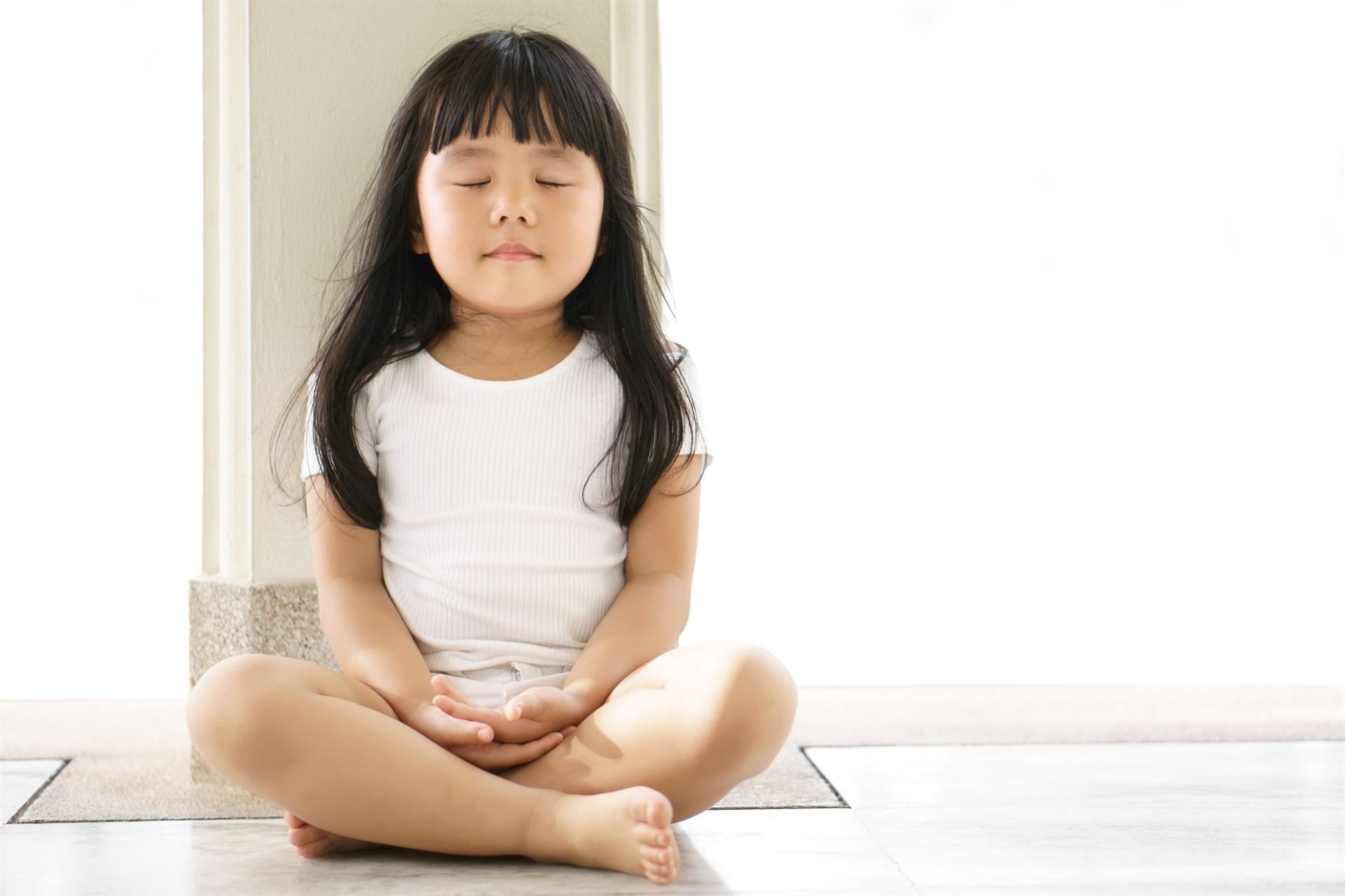 A young girl with long hair sits cross-legged on the floor, eyes closed, appearing calm and meditative.