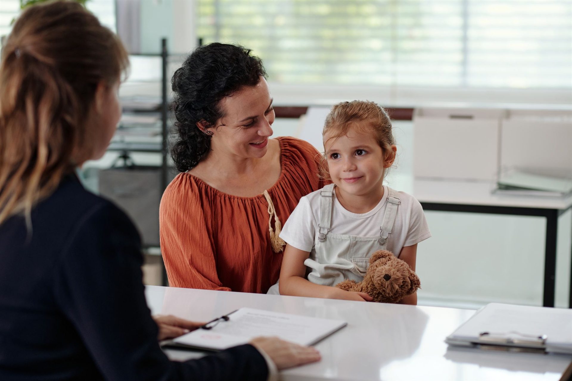 A woman and a girl sit together, smiling at another woman across a table, with documents and a stuffed animal visible.