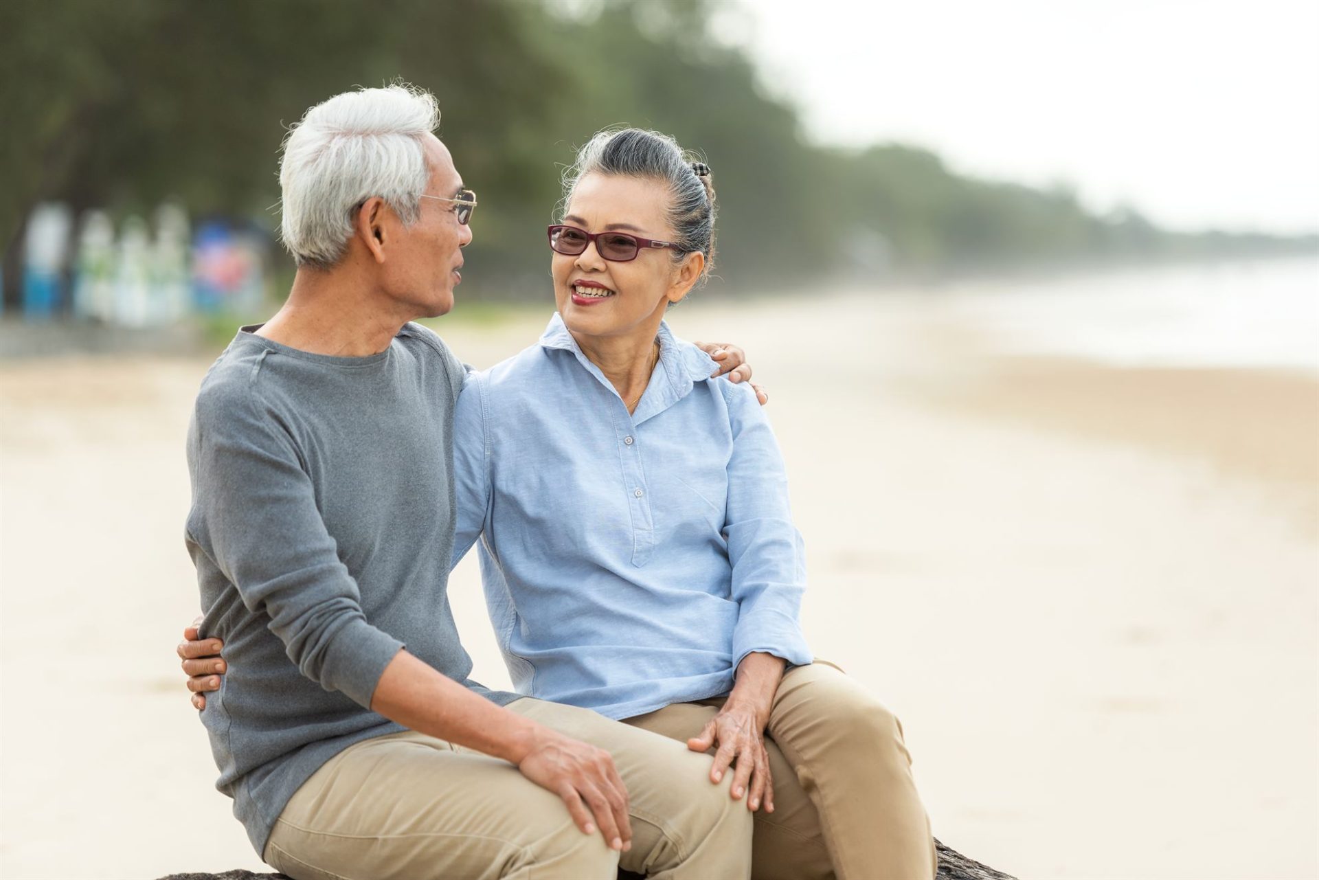 An elderly couple sits on a log at the beach, smiling and engaging with each other.