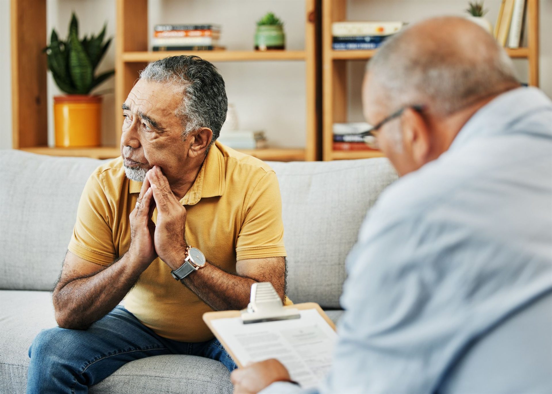 An older man in a yellow shirt sits on a couch, looking thoughtfully at another man holding a clipboard.
