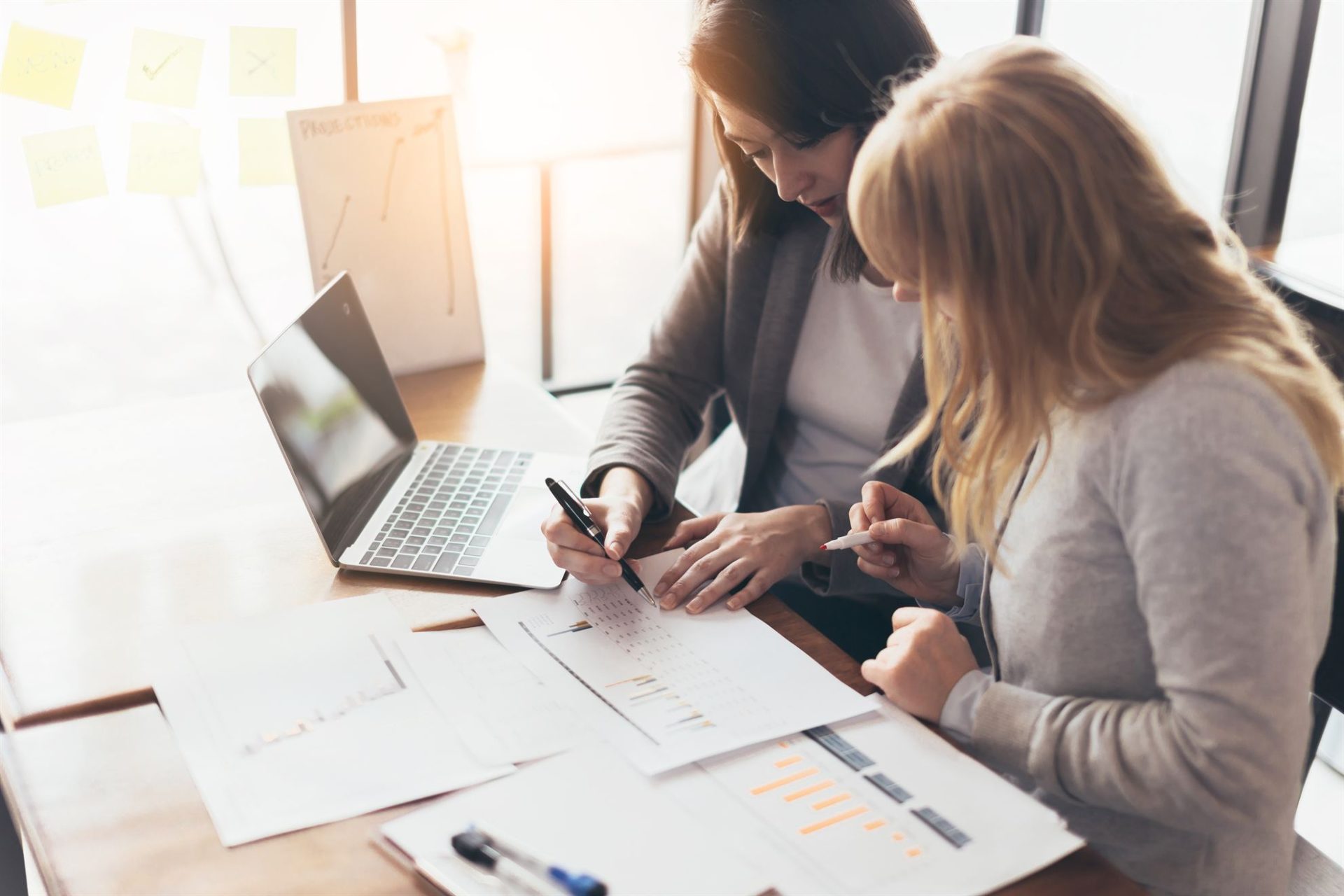Two women collaborate at a table with a laptop, documents, and charts, discussing project details in a bright office.