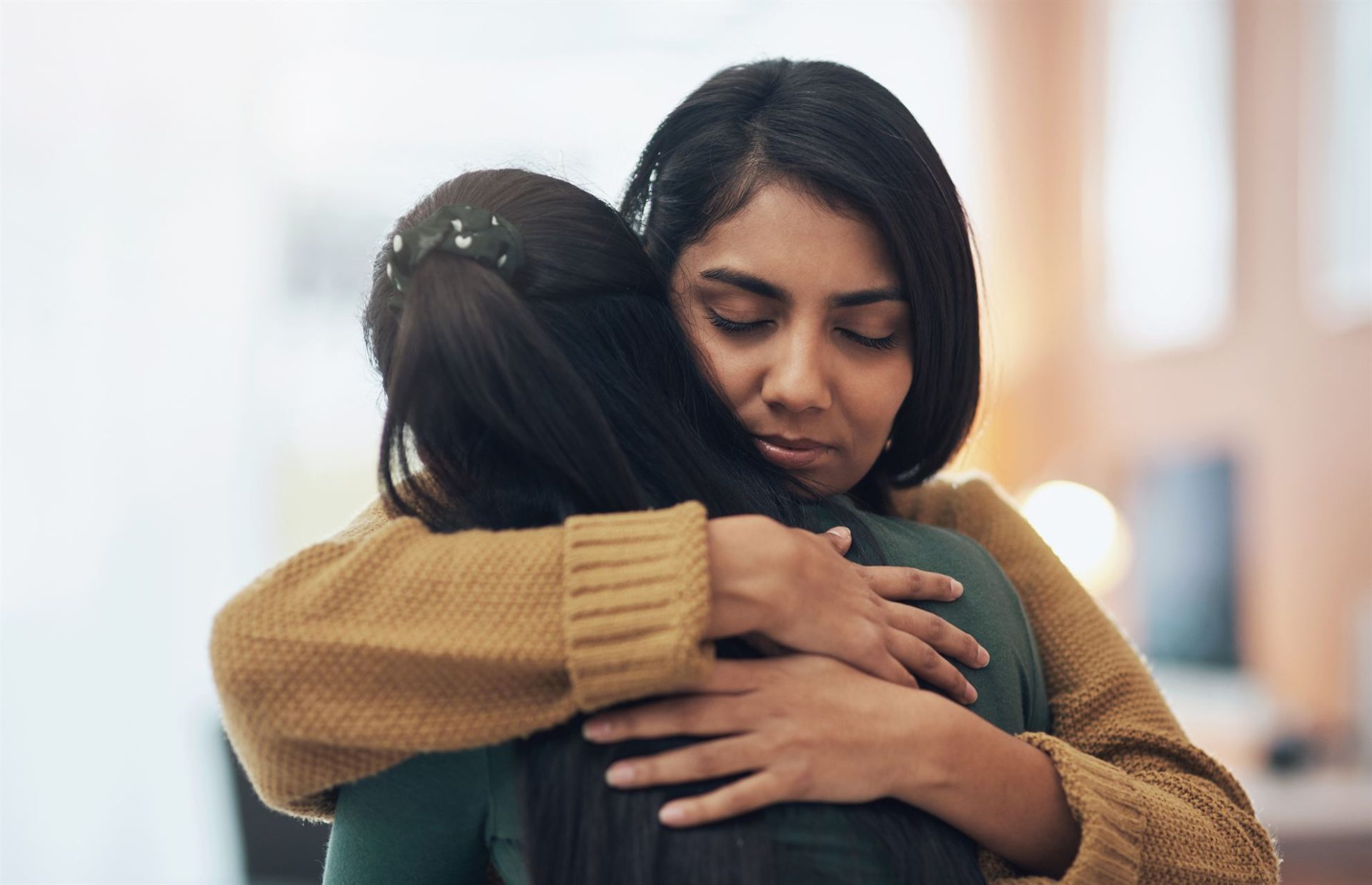 Two women embrace each other, showing comfort and support, in a softly lit indoor setting.