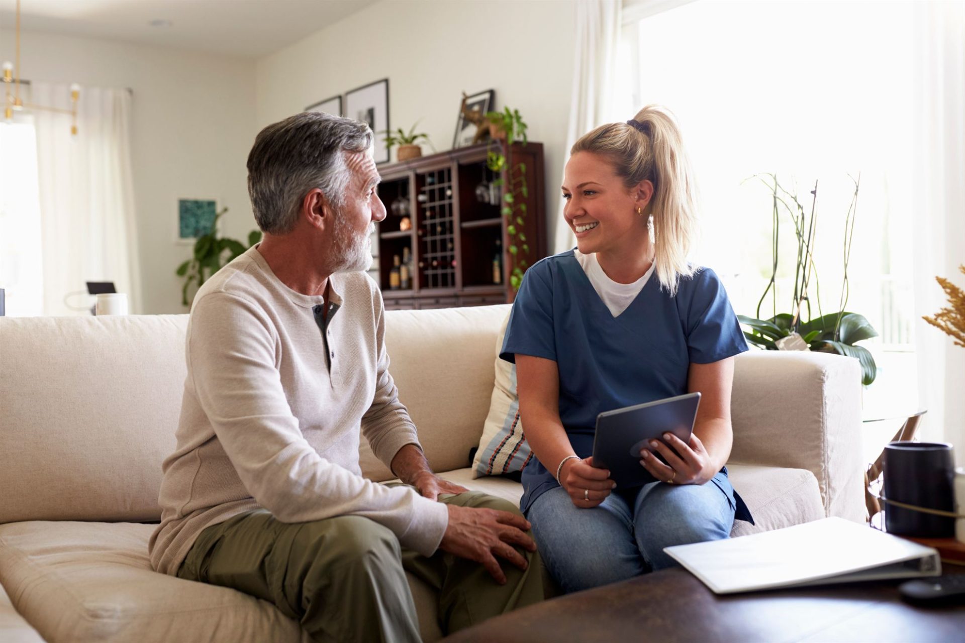 A smiling woman in scrubs talks with an older man on a couch in a bright, cozy living room.