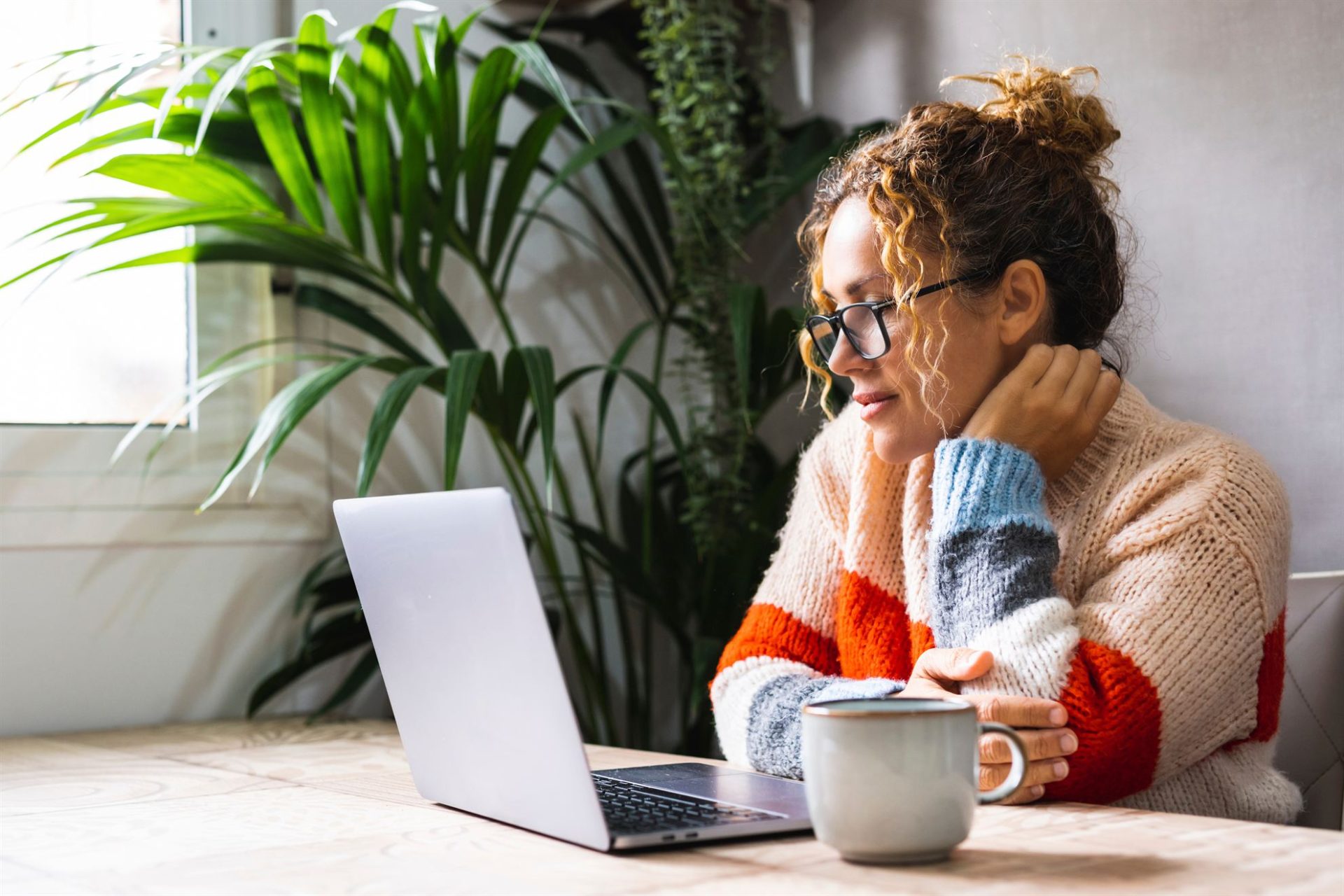 A woman with curly hair, wearing a colorful sweater, sits at a table with a laptop and a mug, surrounded by plants.