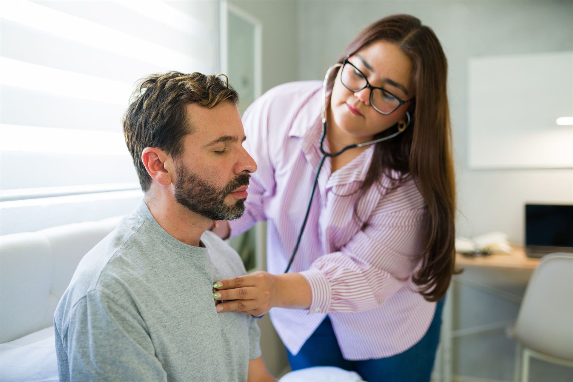 A healthcare professional examines a patient using a stethoscope in a light-filled room.