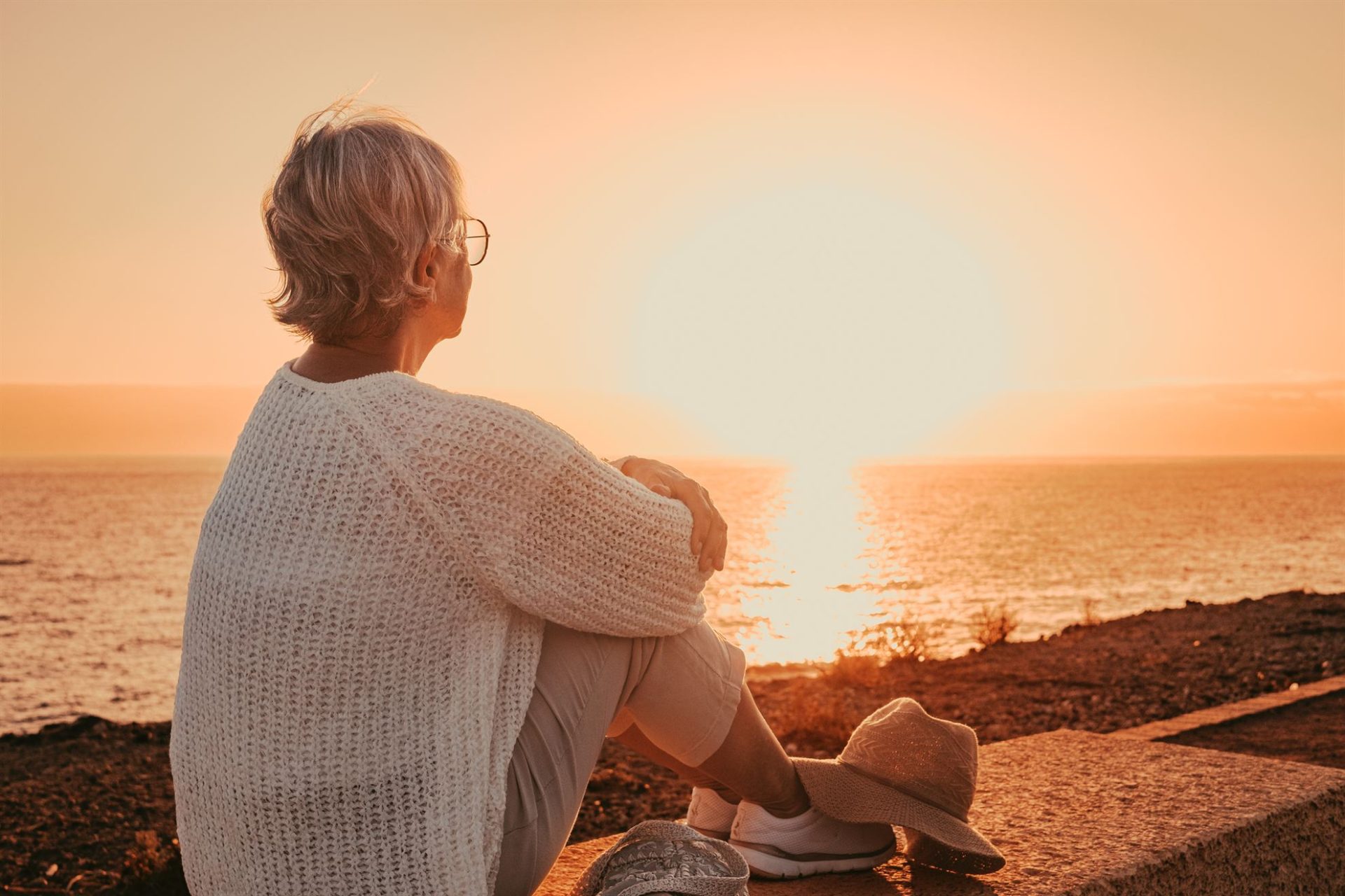 A woman with short hair sits by the ocean, watching a sunset, wearing a white sweater and a hat.