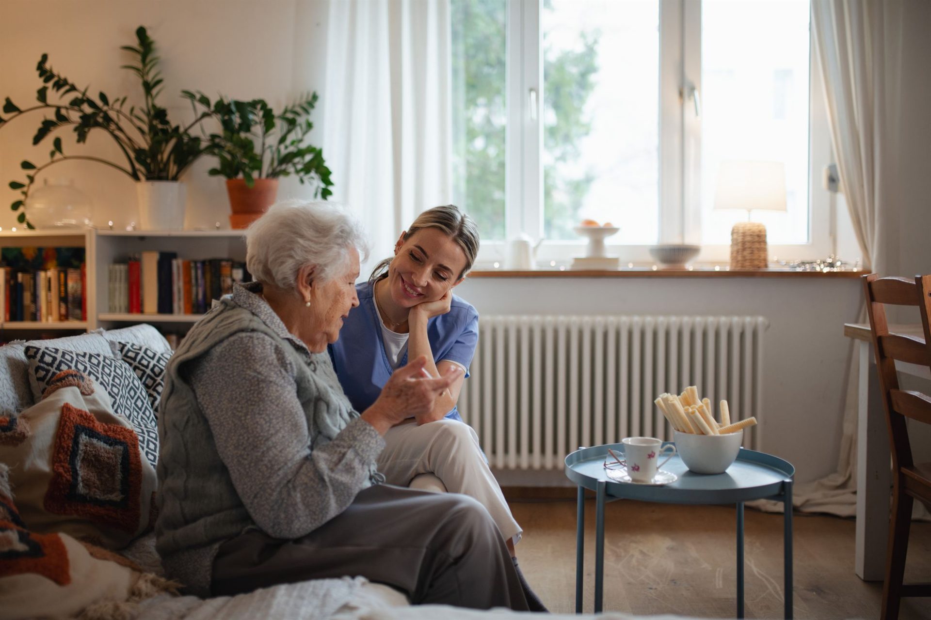 A caregiver and an elderly woman are sitting together, smiling, with snacks and a cup on a small table nearby.