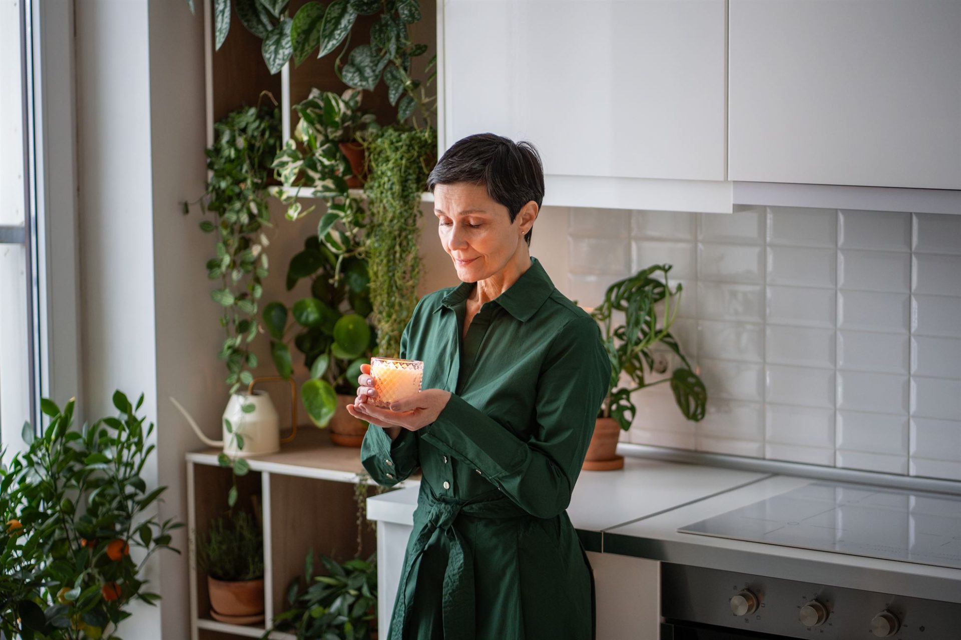 Woman in a green dress holding a lit candle in a kitchen surrounded by indoor plants.