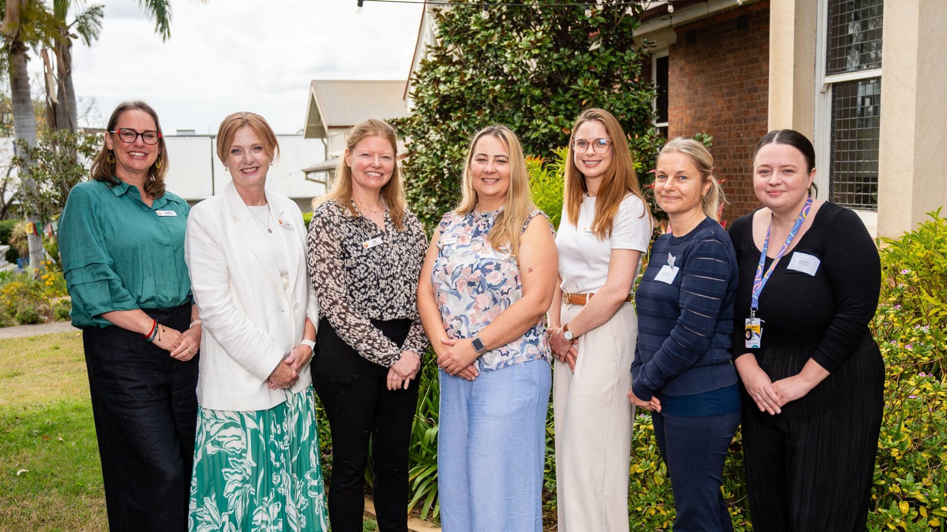 Seven women stand together outdoors, smiling, in front of greenery and a building. They are dressed in various professional outfits.